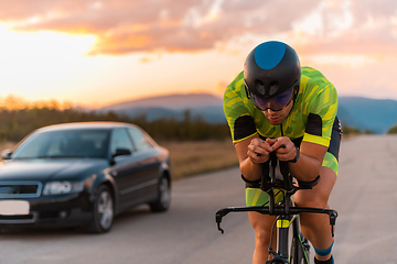 Image showing Close up photo of triathlete riding his bicycle during sunset, preparing for a marathon. The warm colors of the sky provide a beautiful backdrop for his determined and focused effort.