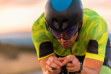 Image showing Close up photo of triathlete riding his bicycle during sunset, preparing for a marathon. The warm colors of the sky provide a beautiful backdrop for his determined and focused effort.