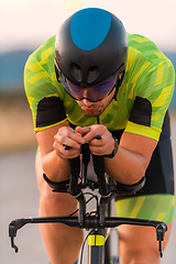 Image showing Close up photo of triathlete riding his bicycle during sunset, preparing for a marathon. The warm colors of the sky provide a beautiful backdrop for his determined and focused effort.