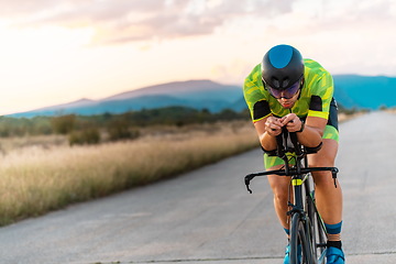 Image showing Triathlete riding his bicycle during sunset, preparing for a marathon. The warm colors of the sky provide a beautiful backdrop for his determined and focused effort.