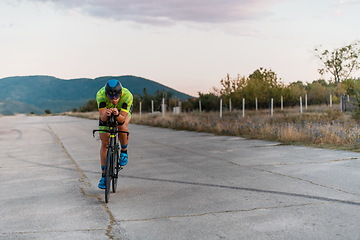 Image showing Triathlete riding his bicycle during sunset, preparing for a marathon. The warm colors of the sky provide a beautiful backdrop for his determined and focused effort.