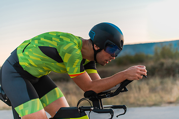 Image showing Close up photo of triathlete riding his bicycle during sunset, preparing for a marathon. The warm colors of the sky provide a beautiful backdrop for his determined and focused effort.