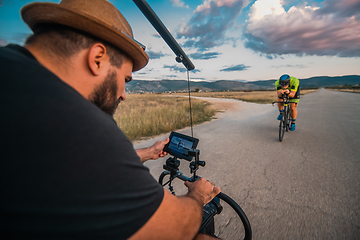 Image showing A videographer recording a triathlete riding his bike preparing for an upcoming marathon.Athlete's physical endurance and the dedication required to succeed in the sport.