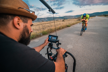 Image showing A videographer recording a triathlete riding his bike preparing for an upcoming marathon.Athlete's physical endurance and the dedication required to succeed in the sport.