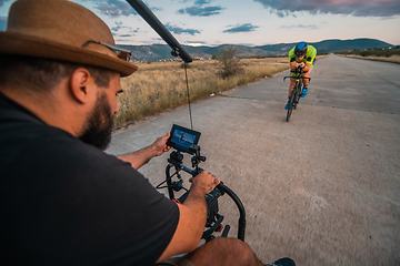 Image showing A videographer recording a triathlete riding his bike preparing for an upcoming marathon.Athlete's physical endurance and the dedication required to succeed in the sport.