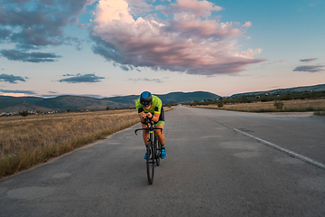 Image showing Triathlete riding his bicycle during sunset, preparing for a marathon. The warm colors of the sky provide a beautiful backdrop for his determined and focused effort.