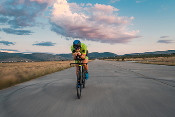 Image showing Triathlete riding his bicycle during sunset, preparing for a marathon. The warm colors of the sky provide a beautiful backdrop for his determined and focused effort.