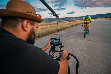 Image showing A videographer recording a triathlete riding his bike preparing for an upcoming marathon.Athlete's physical endurance and the dedication required to succeed in the sport.