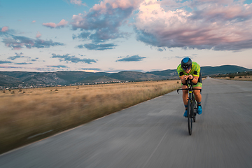 Image showing Triathlete riding his bicycle during sunset, preparing for a marathon. The warm colors of the sky provide a beautiful backdrop for his determined and focused effort.