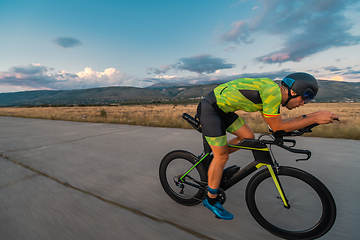 Image showing Triathlete riding his bicycle during sunset, preparing for a marathon. The warm colors of the sky provide a beautiful backdrop for his determined and focused effort.