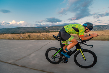 Image showing Triathlete riding his bicycle during sunset, preparing for a marathon. The warm colors of the sky provide a beautiful backdrop for his determined and focused effort.