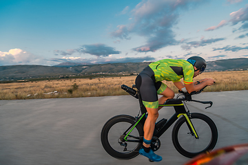 Image showing Triathlete riding his bicycle during sunset, preparing for a marathon. The warm colors of the sky provide a beautiful backdrop for his determined and focused effort.