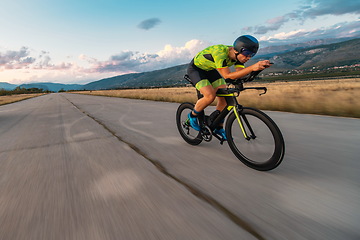 Image showing Triathlete riding his bicycle during sunset, preparing for a marathon. The warm colors of the sky provide a beautiful backdrop for his determined and focused effort.