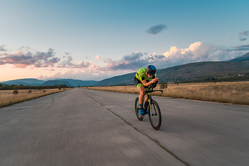 Image showing Triathlete riding his bicycle during sunset, preparing for a marathon. The warm colors of the sky provide a beautiful backdrop for his determined and focused effort.