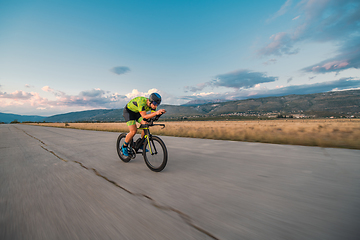 Image showing Triathlete riding his bicycle during sunset, preparing for a marathon. The warm colors of the sky provide a beautiful backdrop for his determined and focused effort.