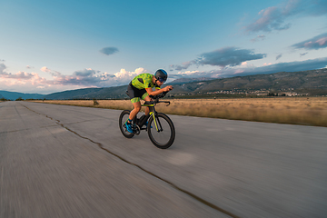 Image showing Triathlete riding his bicycle during sunset, preparing for a marathon. The warm colors of the sky provide a beautiful backdrop for his determined and focused effort.