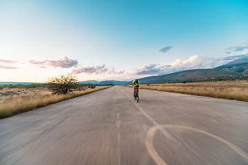 Image showing Triathlete riding his bicycle during sunset, preparing for a marathon. The warm colors of the sky provide a beautiful backdrop for his determined and focused effort.