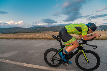 Image showing Triathlete riding his bicycle during sunset, preparing for a marathon. The warm colors of the sky provide a beautiful backdrop for his determined and focused effort.