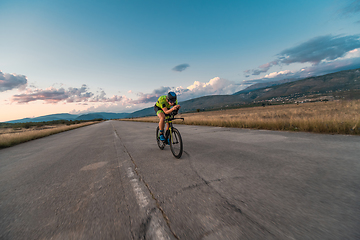 Image showing Triathlete riding his bicycle during sunset, preparing for a marathon. The warm colors of the sky provide a beautiful backdrop for his determined and focused effort.