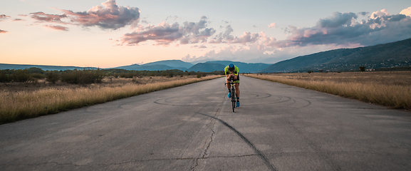 Image showing Triathlete riding his bicycle during sunset, preparing for a marathon. The warm colors of the sky provide a beautiful backdrop for his determined and focused effort.