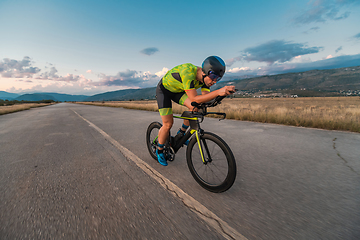 Image showing Triathlete riding his bicycle during sunset, preparing for a marathon. The warm colors of the sky provide a beautiful backdrop for his determined and focused effort.