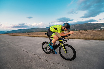 Image showing Triathlete riding his bicycle during sunset, preparing for a marathon. The warm colors of the sky provide a beautiful backdrop for his determined and focused effort.