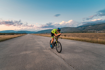 Image showing Triathlete riding his bicycle during sunset, preparing for a marathon. The warm colors of the sky provide a beautiful backdrop for his determined and focused effort.