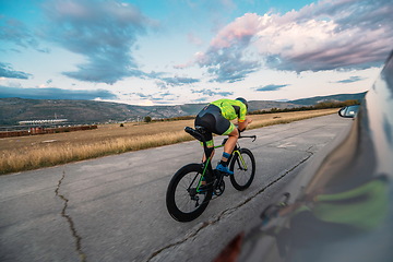 Image showing Triathlete riding his bicycle during sunset, preparing for a marathon. The warm colors of the sky provide a beautiful backdrop for his determined and focused effort.