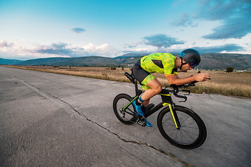 Image showing Triathlete riding his bicycle during sunset, preparing for a marathon. The warm colors of the sky provide a beautiful backdrop for his determined and focused effort.