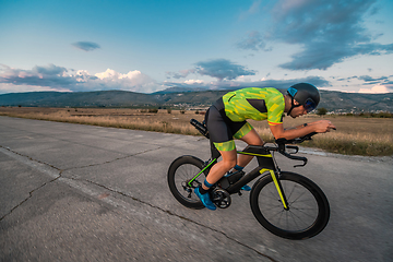 Image showing Triathlete riding his bicycle during sunset, preparing for a marathon. The warm colors of the sky provide a beautiful backdrop for his determined and focused effort.