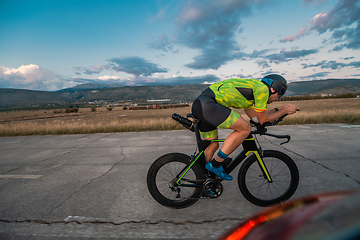 Image showing Triathlete riding his bicycle during sunset, preparing for a marathon. The warm colors of the sky provide a beautiful backdrop for his determined and focused effort.