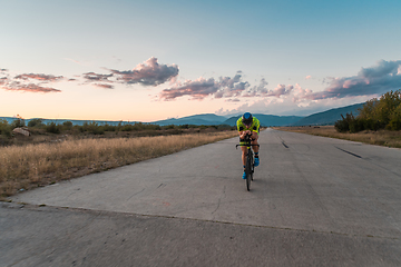 Image showing Triathlete riding his bicycle during sunset, preparing for a marathon. The warm colors of the sky provide a beautiful backdrop for his determined and focused effort.