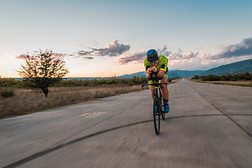 Image showing Triathlete riding his bicycle during sunset, preparing for a marathon. The warm colors of the sky provide a beautiful backdrop for his determined and focused effort.