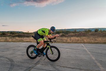 Image showing Triathlete riding his bicycle during sunset, preparing for a marathon. The warm colors of the sky provide a beautiful backdrop for his determined and focused effort.