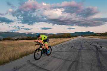 Image showing Triathlete riding his bicycle during sunset, preparing for a marathon. The warm colors of the sky provide a beautiful backdrop for his determined and focused effort.