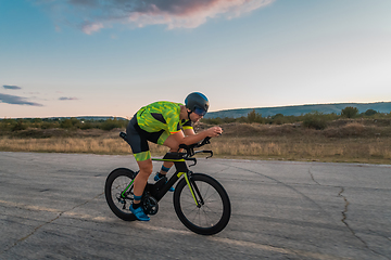 Image showing Triathlete riding his bicycle during sunset, preparing for a marathon. The warm colors of the sky provide a beautiful backdrop for his determined and focused effort.