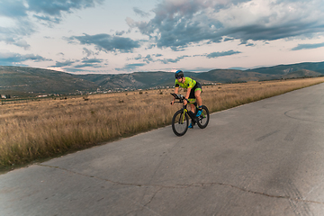 Image showing Triathlete riding his bicycle during sunset, preparing for a marathon. The warm colors of the sky provide a beautiful backdrop for his determined and focused effort.