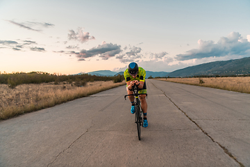 Image showing Triathlete riding his bicycle during sunset, preparing for a marathon. The warm colors of the sky provide a beautiful backdrop for his determined and focused effort.