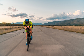 Image showing Triathlete riding his bicycle during sunset, preparing for a marathon. The warm colors of the sky provide a beautiful backdrop for his determined and focused effort.