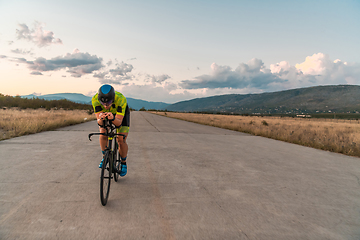 Image showing Triathlete riding his bicycle during sunset, preparing for a marathon. The warm colors of the sky provide a beautiful backdrop for his determined and focused effort.