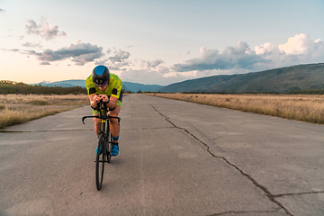 Image showing Triathlete riding his bicycle during sunset, preparing for a marathon. The warm colors of the sky provide a beautiful backdrop for his determined and focused effort.