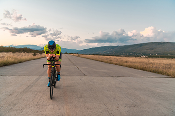 Image showing Triathlete riding his bicycle during sunset, preparing for a marathon. The warm colors of the sky provide a beautiful backdrop for his determined and focused effort.