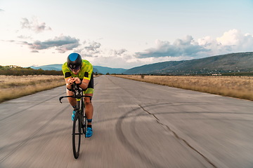 Image showing Triathlete riding his bicycle during sunset, preparing for a marathon. The warm colors of the sky provide a beautiful backdrop for his determined and focused effort.