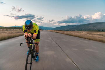 Image showing Triathlete riding his bicycle during sunset, preparing for a marathon. The warm colors of the sky provide a beautiful backdrop for his determined and focused effort.