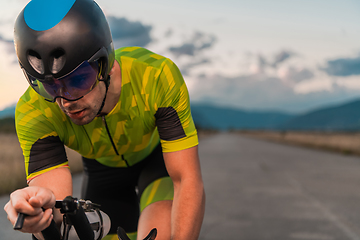 Image showing Close up photo of triathlete riding his bicycle during sunset, preparing for a marathon. The warm colors of the sky provide a beautiful backdrop for his determined and focused effort.