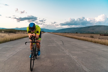 Image showing Triathlete riding his bicycle during sunset, preparing for a marathon. The warm colors of the sky provide a beautiful backdrop for his determined and focused effort.