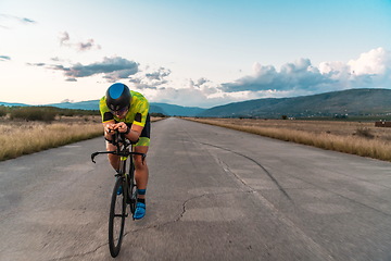 Image showing Triathlete riding his bicycle during sunset, preparing for a marathon. The warm colors of the sky provide a beautiful backdrop for his determined and focused effort.