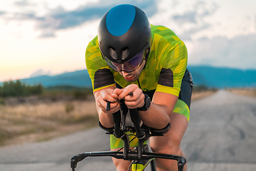 Image showing Close up photo of triathlete riding his bicycle during sunset, preparing for a marathon. The warm colors of the sky provide a beautiful backdrop for his determined and focused effort.