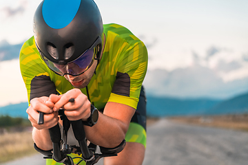 Image showing Close up photo of triathlete riding his bicycle during sunset, preparing for a marathon. The warm colors of the sky provide a beautiful backdrop for his determined and focused effort.