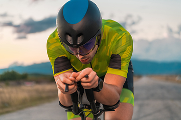 Image showing Close up photo of triathlete riding his bicycle during sunset, preparing for a marathon. The warm colors of the sky provide a beautiful backdrop for his determined and focused effort.